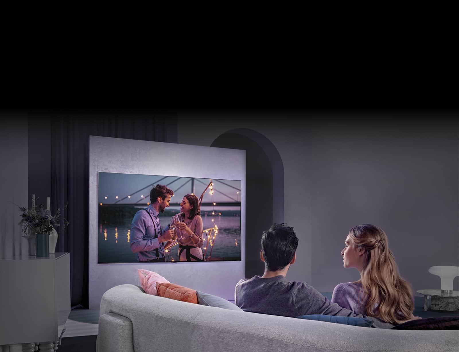 Osoba siedząca na kanapie i oglądająca film na dużym telewizorze na ścianie.
